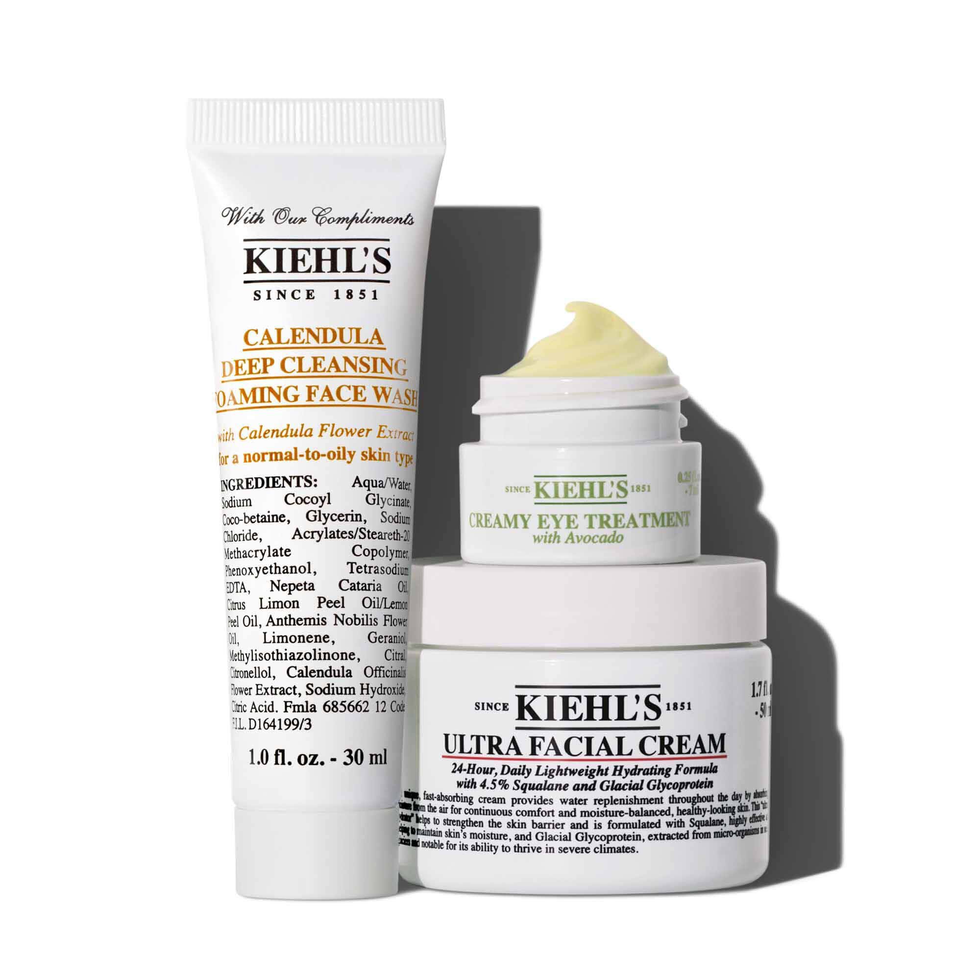 Koyu yeşil bir zemin uzerinde parlak yeşil detaylarla tasarlanmış bir grafik, Kiehl’s hediye kutusunu ve beyaz ambalajında uc cilt bakım urununu one cıkarıyor: "KIEHL’S CALENDULA DEEP CLEANSING FOAMING FACE WASH" tupu, urunun gorunduğu "KIEHL’S CREAMY EYE TREATMENT WITH AVOCADO" kavanozu ve "KIEHL’S ULTRA FACIAL CREAM" kavanozu. Sağda, yeşil bir “K” harfine sahip altın bir sus yer alıyor. Parlak yeşil etikette şu ifade yazıyor: "Bu tatilde YATIŞTIRICI CİLT BAKIMINI hediye edin!" Stilize edilmiş parlak yeşil bir kurdele grafiği hediye kutusunu susluyor.