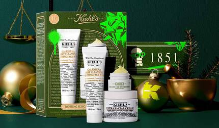 Kiehl’s hediye seti, altın suslemeler, altın terazi ve yeşil dallarla suslenmiş şenlikli bir yeşil arka plan onunde sergileniyor. Set, yeşil ve altın detaylara, neon yeşil aksanlara ve Kiehl’s logosuna sahip bir kutunun onunde duzenlenmiş uc urunden oluşuyor: Calendula Deep Cleansing Foaming Face Wash, Creamy Eye Treatment with Avocado ve Ultra Facial Cream. Fon kısmında, '1851' yazısını ve bir kafatası tasarımını iceren yeşil bir kutu gorunerek yılbaşı temasını tamamlıyor.