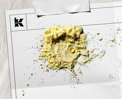 Yakından cekilmiş bir goruntude, beyaz bir kağıt uzerinde acık sarı renkte bir toz yığını yer alıyor. Tozun icinde kucuk, berrak bir sıvı damlası goruluyor. Kağıdın sol ust koşesinde bir 'K' logosu bulunuyor ve kağıt hafifce buruşmuş, ust kısmından bantlanmış gorunuyor.