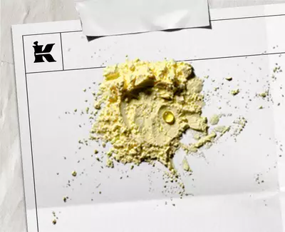 Yakından cekilmiş bir goruntude, beyaz bir kağıt uzerinde acık sarı renkte bir toz yığını yer alıyor. Tozun icinde kucuk, berrak bir sıvı damlası goruluyor. Kağıdın sol ust koşesinde bir 'K' logosu bulunuyor ve kağıt hafifce buruşmuş, ust kısmından bantlanmış gorunuyor.