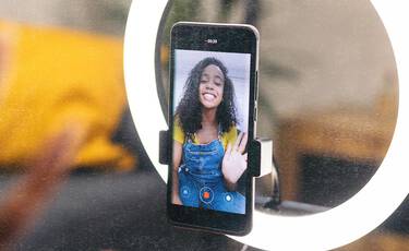 Smartphone mounted in a ring light showing a young woman waving on screen, depicting digital content creation.