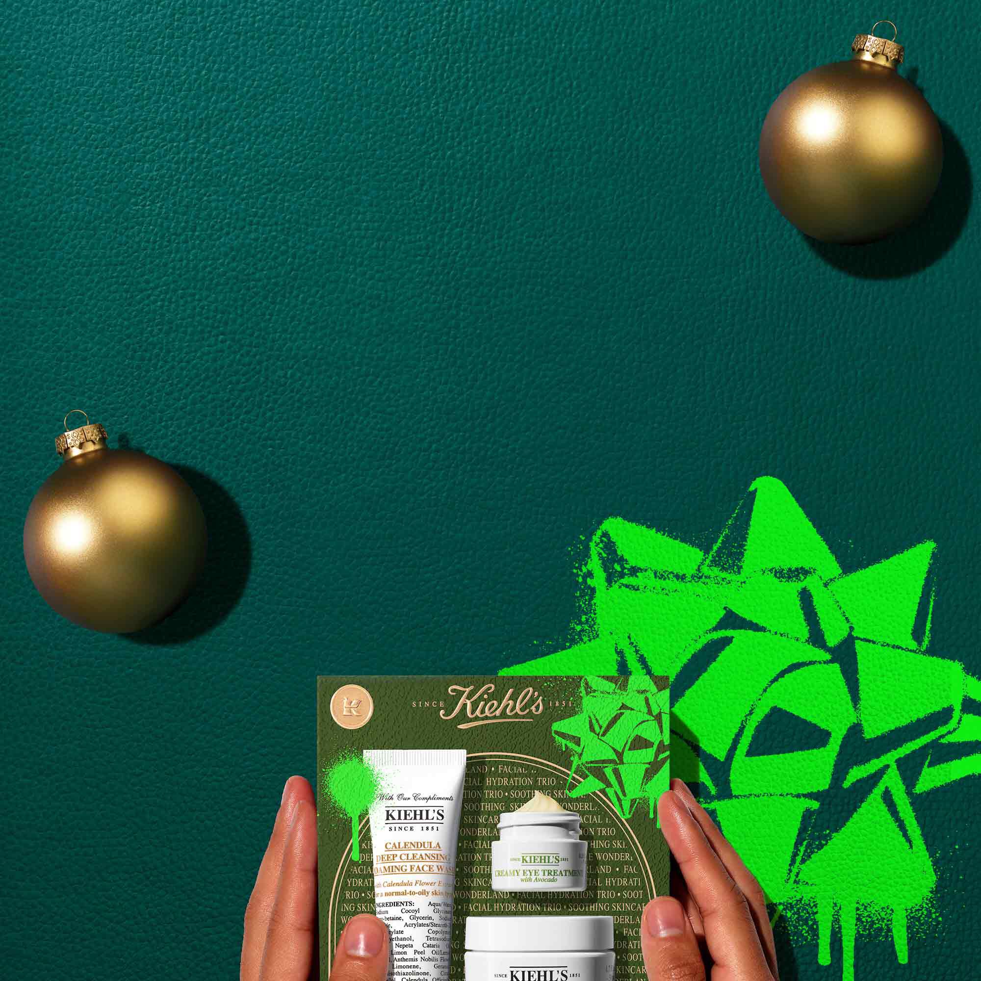 A graphic set against a dark green textured background with bright green graphic accents features three Kiehl’s skincare products: a tube of "KIEHL’S CALENDULA DEEP CLEANSING FOAMING FACE WASH," a jar of "KIEHL’S CREAMY EYE TREATMENT WITH AVOCADO" with visible product, and a jar of "KIEHL’S ULTRA FACIAL CREAM." Two gold ornaments are placed on either side of the products. Below, a Kiehl’s gift box displays the same three products, decorated with bright green graphic shapes and a bow design.